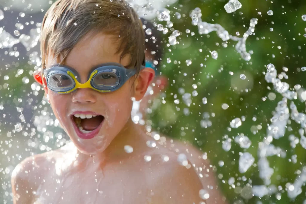 Ein lachendes Kind mit Schwimmbrille spielt fröhlich im spritzenden Wasser.